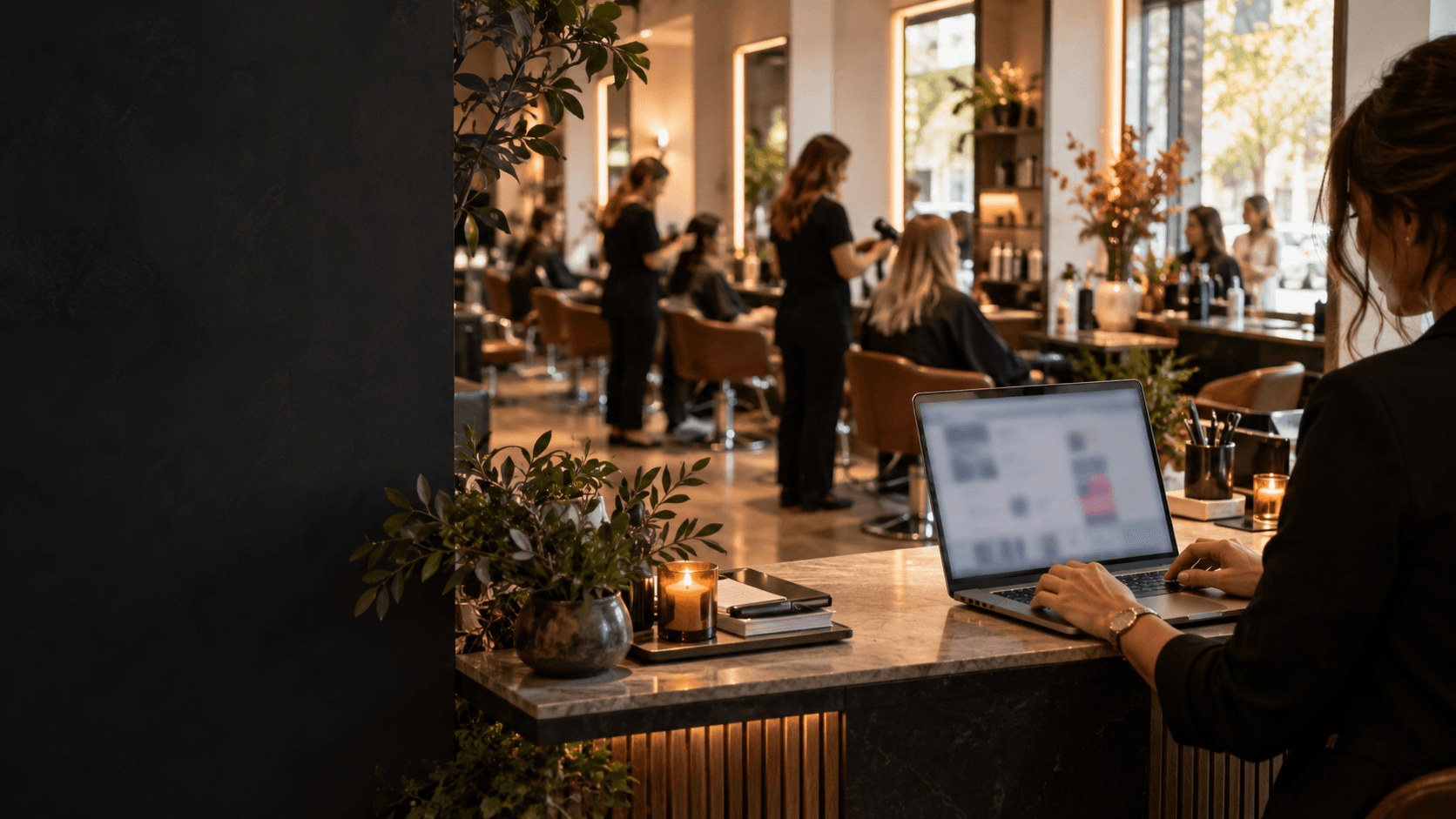 Salon manager using a laptop at a reception desk while stylists work in a bright modern salon